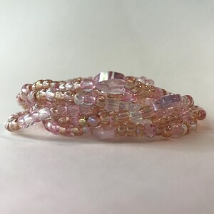 May include: A coiled bracelet composed of small, iridescent beads in shades of pink, peach, and clear. The bracelet features a mix of round and oblong beads, creating a textured and layered appearance. The beads are strung together, forming a flexible accessory.