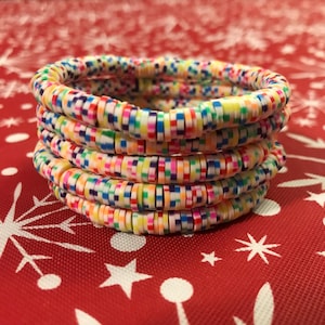 May include: Five colourful bracelets stacked on top of each other. Each bracelet is made of small, flat, rectangular beads in various colours, including red, blue, yellow, green, and pink. The bracelets are on a red surface with white snowflake and star patterns.