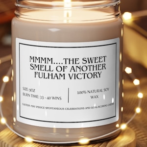 May include: A lit candle in a clear glass jar with a label that reads "MMMM....THE SWEET SMELL OF ANOTHER FULHAM VICTORY." The candle sits on a wooden coaster. The candle is 9oz and has a burn time of 33-40 wins.
