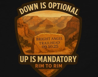 Op de afbeelding: Zwart t-shirt met een afbeelding van een woestijnlandschap en de tekst "DOWN IS OPTIONAL, UP IS MANDATORY, RIM TO RIM." De afbeelding bevat ook "BRIGHT ANGEL TRAILHEAD 09.10.25."