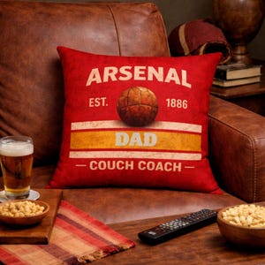May include: Red decorative pillow with the text "ARSENAL" and "DAD - COUCH COACH -" in white. A vintage-style soccer ball graphic is in the center. The pillow is on a brown leather couch.