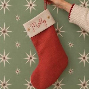 Peut inclure: Une chaussette de Noël rouge avec un revers beige portant le prénom Molly brodé en écriture rouge, encadré par deux petites étoiles dorées. La chaussette comporte une boucle en tissu rouge assortie sur le dessus pour la suspendre.