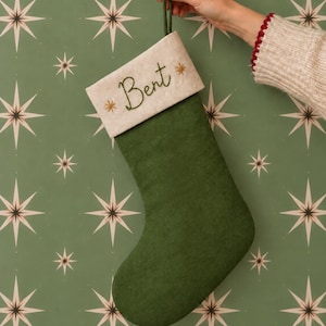 May include: A dark green Christmas stocking with a cream-colored cuff. The cuff features the embroidered name "Bent" in dark green cursive script, flanked by two small, gold-colored embroidered starburst designs. A green fabric loop is attached.