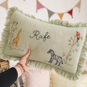 May include: A rectangular sage green cushion with a ruffled edge, featuring embroidered illustrations of a giraffe, a zebra, and a parrot on a branch. The name Rafe is embroidered in dark green cursive script in the centre of the cushion.