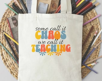 Può includere: Borsa tote color crema con la scritta "some call it CHAOS we call it TEACHING" in stile retrò. La borsa è piena di penne e matite, appoggiata su un cestino intrecciato.