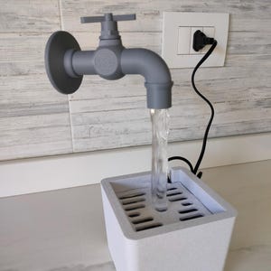Può includere: Una scultura di rubinetto grigio con acqua che scorre in un bacino rettangolare bianco. Il rubinetto è montato su un muro e collegato a una presa di corrente. La scultura è un oggetto decorativo.