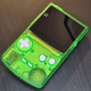 May include: A transparent green handheld gaming console with a black screen. The console has white buttons, a directional pad, and a power button. The word "POWER" is printed on the top of the screen.
