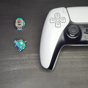 May include: A white and black video game controller with a green and blue joystick replacement. The controller has a white faceplate and a black backplate. The joystick replacement is a small, round device with a green top and a blue base.