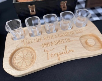 May include: A light-colored wooden serving board with five clear glass shot glasses. The board features engraved text that reads, "Take life with a grain of salt and a shot of Tequila," along with a carved citrus slice icon and a circular indentation.