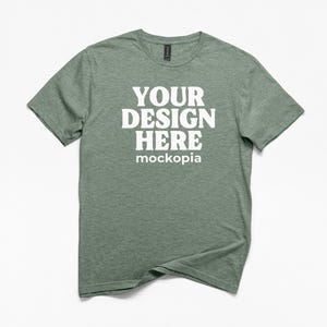 May include: A heather green t-shirt with the text "YOUR DESIGN HERE" in bold white letters. The word "mockopia" is below the main text. The shirt has a crew neck and short sleeves.