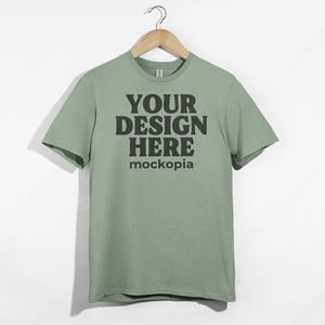 May include: A sage green t-shirt with short sleeves hanging on a wooden hanger. The shirt has the text "YOUR DESIGN HERE" and "mockopia" printed on the front.