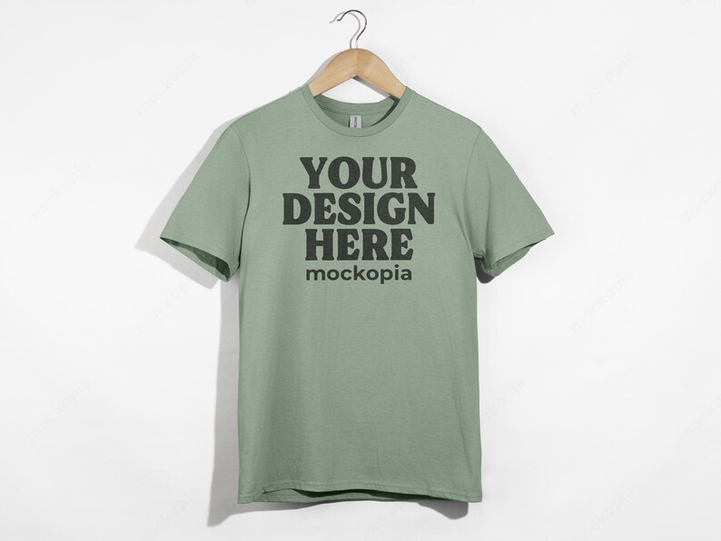 May include: A sage green t-shirt with short sleeves hanging on a wooden hanger. The shirt has the text "YOUR DESIGN HERE" and "mockopia" printed on the front.