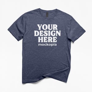 May include: A navy blue t-shirt with the text "YOUR DESIGN HERE mockopia" in white block letters. The shirt has short sleeves and a crew neck. The fabric has a heathered texture.