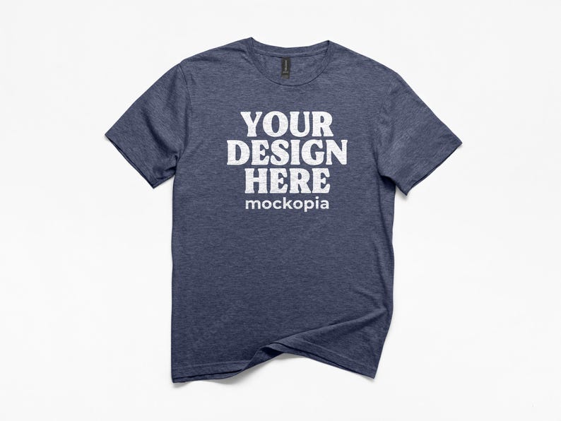 May include: A navy blue t-shirt with the text "YOUR DESIGN HERE mockopia" in white block letters. The shirt has short sleeves and a crew neck. The fabric has a heathered texture.
