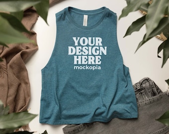 May include: Teal sleeveless crop top with the text "YOUR DESIGN HERE mockopia" in white. The top is made of a soft material and is displayed with a pair of gray shorts and a brown scarf. Green leaves frame the top.