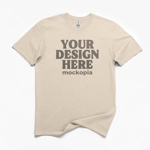 May include: A beige t-shirt with the text "YOUR DESIGN HERE" in a dark brown font. The word "mockopia" is below the text. The t-shirt is displayed on a white background.
