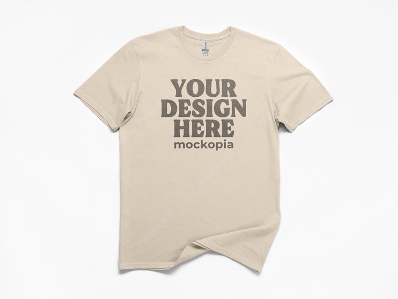 May include: A beige t-shirt with the text "YOUR DESIGN HERE" in a dark brown font. The word "mockopia" is below the text. The t-shirt is displayed on a white background.