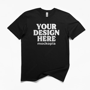 May include: Black crew neck t-shirt with the text "YOUR DESIGN HERE" in a bold, white font. The word "mockopia" is printed below the main text. The t-shirt is displayed on a white background.