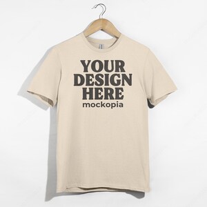 May include: A beige t-shirt with short sleeves, hanging on a wooden hanger. The shirt has the text "YOUR DESIGN HERE" and "mockopia" printed on the front in black.
