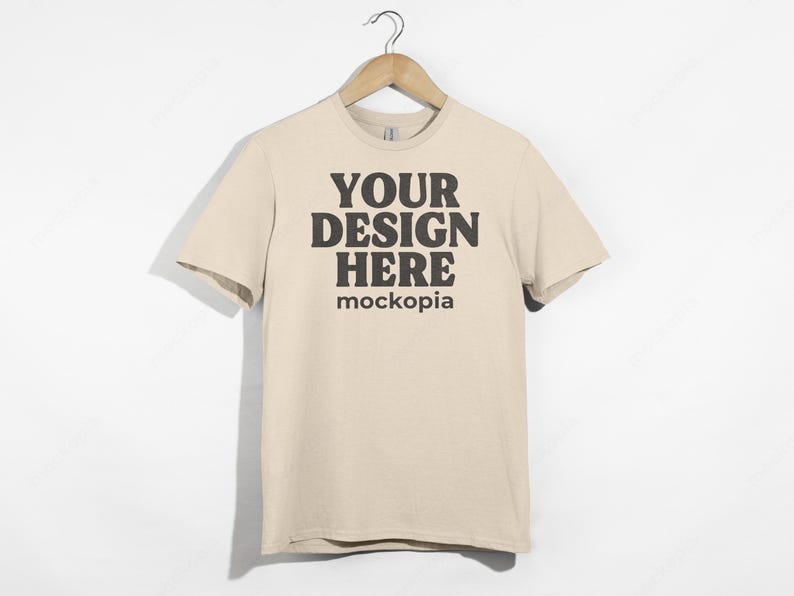 May include: A beige t-shirt with short sleeves, hanging on a wooden hanger. The shirt has the text "YOUR DESIGN HERE" and "mockopia" printed on the front in black.