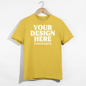 May include: A yellow t-shirt with short sleeves hanging on a wooden hanger. The shirt has the text "YOUR DESIGN HERE" and "mockopia" printed on the front in white.