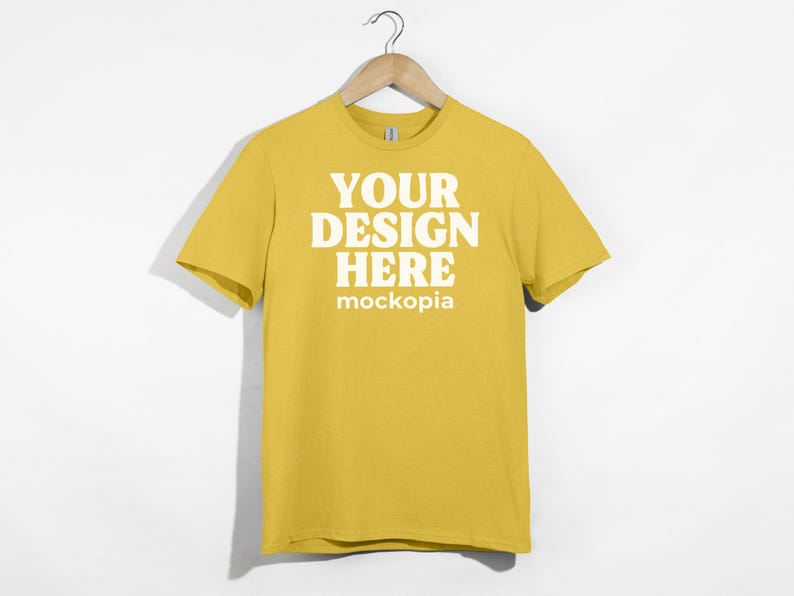 May include: A yellow t-shirt with short sleeves hanging on a wooden hanger. The shirt has the text "YOUR DESIGN HERE" and "mockopia" printed on the front in white.