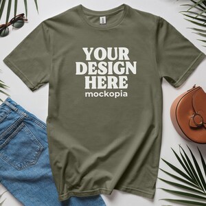 Op de afbeelding: Een groen T-shirt met de tekst "YOUR DESIGN HERE mockopia" gedrukt op de voorkant. Het T-shirt ligt plat op een wit oppervlak met een blauwe spijkerbroek, een zonnebril en een bruine handtas.