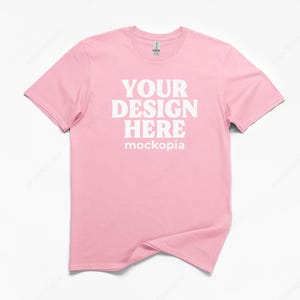 May include: A light pink t-shirt with the text "YOUR DESIGN HERE" in white, with the word "mockopia" below. The shirt is a crew neck style and is displayed on a white background.