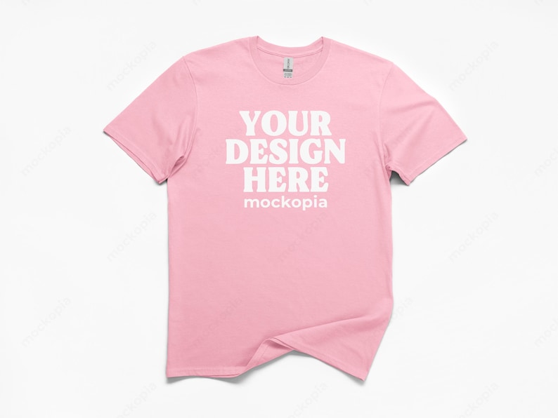 May include: A light pink t-shirt with the text "YOUR DESIGN HERE" in white, with the word "mockopia" below. The shirt is a crew neck style and is displayed on a white background.