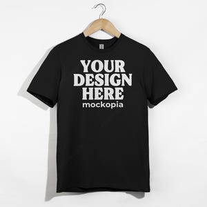 Op de afbeelding: Een zwart t-shirt met de tekst "YOUR DESIGN HERE" in grote witte hoofdletters, met "mockopia" in kleinere witte letters eronder. Het t-shirt hangt aan een houten hanger tegen een witte achtergrond.