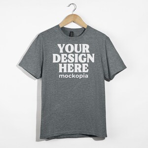 May include: A heather gray t-shirt with short sleeves and a crew neck. The shirt has the text "YOUR DESIGN HERE" and "mockopia" printed on the front in white.