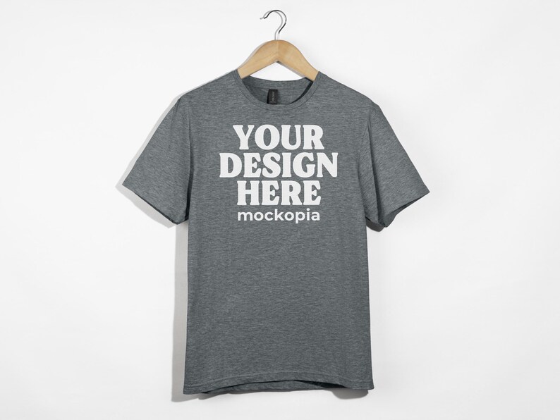 May include: A heather gray t-shirt with short sleeves and a crew neck. The shirt has the text "YOUR DESIGN HERE" and "mockopia" printed on the front in white.