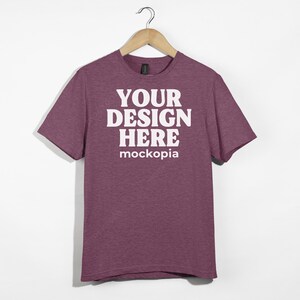 May include: A maroon heather t-shirt with short sleeves, hanging on a wooden hanger. The shirt has the text "YOUR DESIGN HERE" and "mockopia" printed on the front in white.