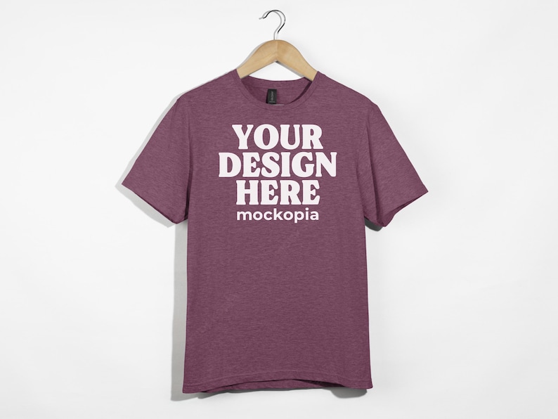 May include: A maroon heather t-shirt with short sleeves, hanging on a wooden hanger. The shirt has the text "YOUR DESIGN HERE" and "mockopia" printed on the front in white.