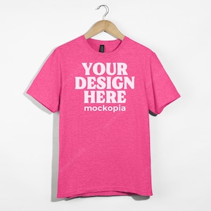 May include: Pink t-shirt with short sleeves and a crew neck. The shirt has the text "YOUR DESIGN HERE" and "mockopia" printed on the front.