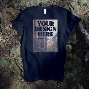 Può includere: Una maglietta blu scuro con una stampa bianca che dice "YOUR DESIGN HERE mockopia". La maglietta è sdraiata su un letto di muschio e ramoscelli.