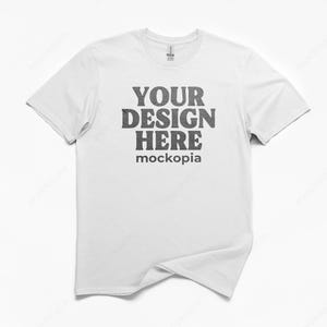 May include: A white t-shirt with a crew neck. The front of the shirt features the text "YOUR DESIGN HERE" in a dark gray, distressed font, with "mockopia" below it. The shirt is displayed on a white background.