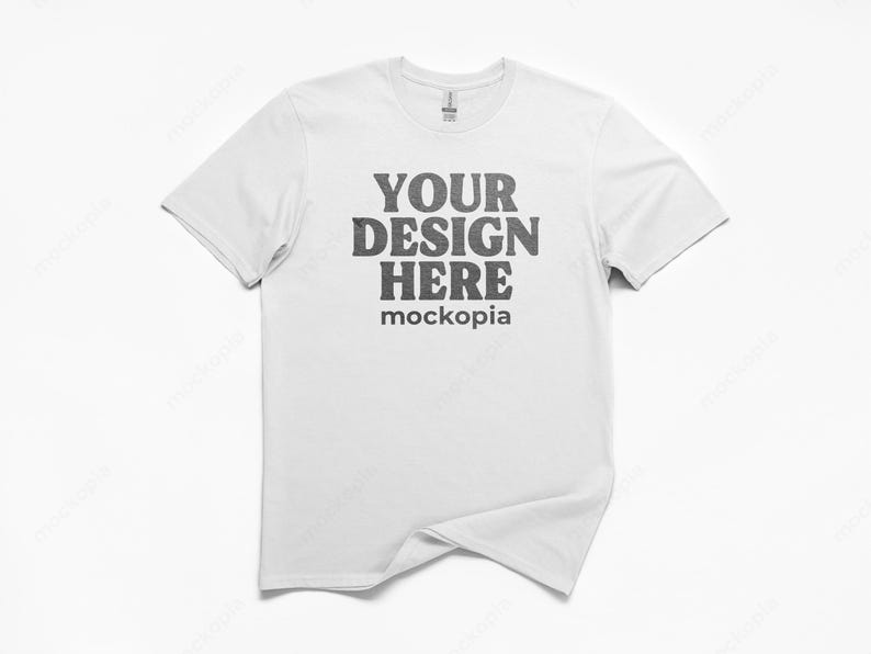 May include: A white t-shirt with a crew neck. The front of the shirt features the text "YOUR DESIGN HERE" in a dark gray, distressed font, with "mockopia" below it. The shirt is displayed on a white background.