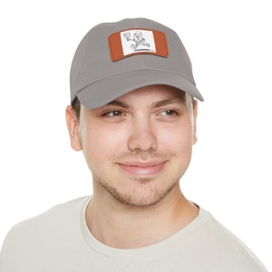 May include: A grey baseball cap with a brown leather patch showing a cartoon character with a camera. The cap has a curved peak and a classic six-panel design.