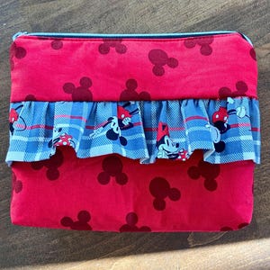 May include: Red zippered pouch with a Mickey Mouse theme. The pouch features a red fabric with Mickey Mouse silhouettes and a blue and white plaid ruffle with Mickey and Minnie Mouse characters.