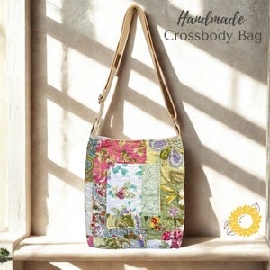 May include: A quilted crossbody bag featuring a patchwork design with various floral patterns in shades of pink, green, yellow, and white. The bag includes a long, adjustable beige strap and the text "Handmade Crossbody Bag" is visible above.