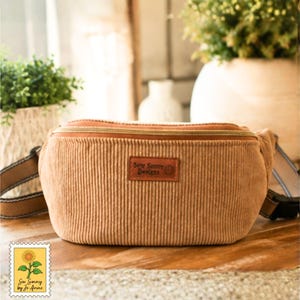 May include: A tan corduroy fanny pack featuring a horizontal zipper closure and a rectangular brown leather patch centered on the front. The patch is embossed with the text Sew Sunny Designs. The bag includes an adjustable strap with a black buckle.