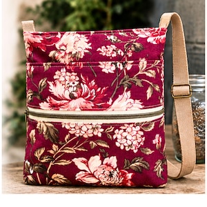 May include: A rectangular crossbody bag featuring a deep burgundy fabric with a prominent floral pattern in shades of pink and white. The bag includes a front zippered pocket with a cream-colored trim and an adjustable beige woven shoulder strap.