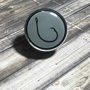 May include: A round, gray, resin cabochon with a black fishing hook design.
