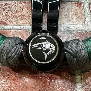 May include: Black steering wheel knob with a green and gray paracord wrap. The knob features a silver fish design on a black background.