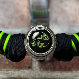 May include: A black and green paracord bracelet with a silver metal charm featuring a green fish design.
