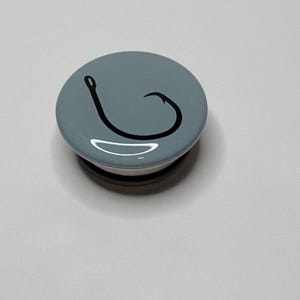May include: A circular phone grip with a smooth, glossy light blue surface featuring a black graphic of a single fishing hook centered on the top. The accessory has a rounded profile and a compact design, providing a functional grip for a mobile device.