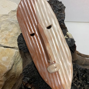 Op de afbeelding: Een keramiek masker met een bruin en wit gestreept ontwerp. Het masker heeft een gestileerd gezicht met een lange neus en gesloten ogen.