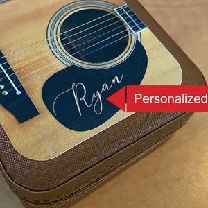 May include: A brown guitar case with a design resembling an acoustic guitar. The case features a black pickguard with the name "Ryan" in cursive. The text "Personalized!" is in a red arrow. The case has a brown zipper and trim.