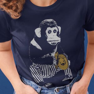 May include: Navy blue t-shirt featuring a vintage-style illustration of a monkey wearing striped pants and holding a yellow hat. The monkey graphic is in shades of white and navy. The shirt is a crew neck style.